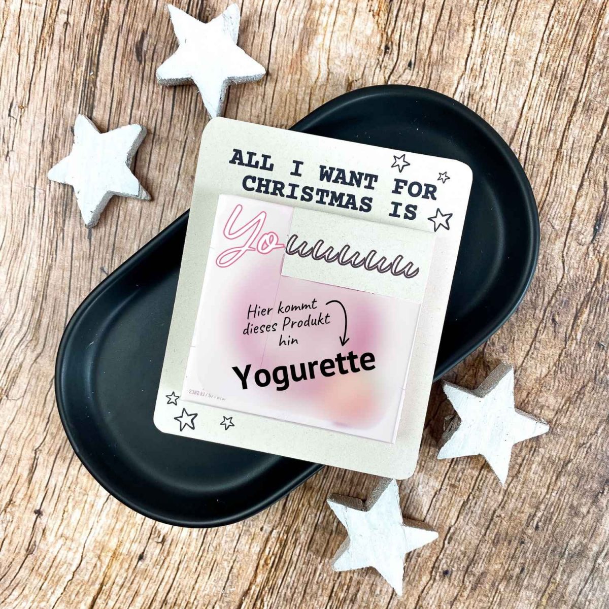 Karte mit Aufschrift „All I want for Christmas is You“ und Platz für eine Yogurette-Schokolade. Liegt auf schwarzer Schale mit weißen Holzsternen. Liebevolle Druckvorlage für ein kleines Weihnachtsgeschenk zum Selbermachen.