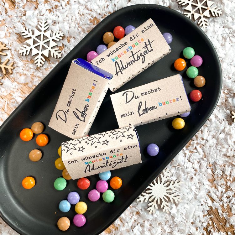Drei kleine Smartiesverpackungen auf schwarzer Schale mit Aufschrift „Du machst mein Leben bunter“ und „Ich wünsche dir eine kunterbunte Adventszeit“. Umgeben von bunten Schokolinsen und Schneeflocken-Deko. Kleine Geschenkidee zu Weihnachten zum Ausdrucken.