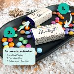Schritt-für-Schritt-Bild zeigt zwei Smarties-Verpackungen mit Aufschriften „Du machst mein Leben bunter“ und „Ich wünsche dir eine kunterbunte Adventszeit“. Umgeben von bunten Smarties und Holzschneeflocken – kreative Geschenkidee zum Ausdrucken.