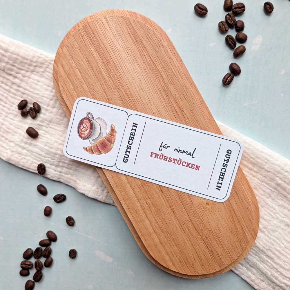 Gutscheinvorlage „Frühstücken“ mit Kaffee- und Croissant-Illustration auf Holzbrett dekoriert. Zeit statt Zeug Geschenkidee zum Ausdrucken für ein gemeinsames Frühstück.