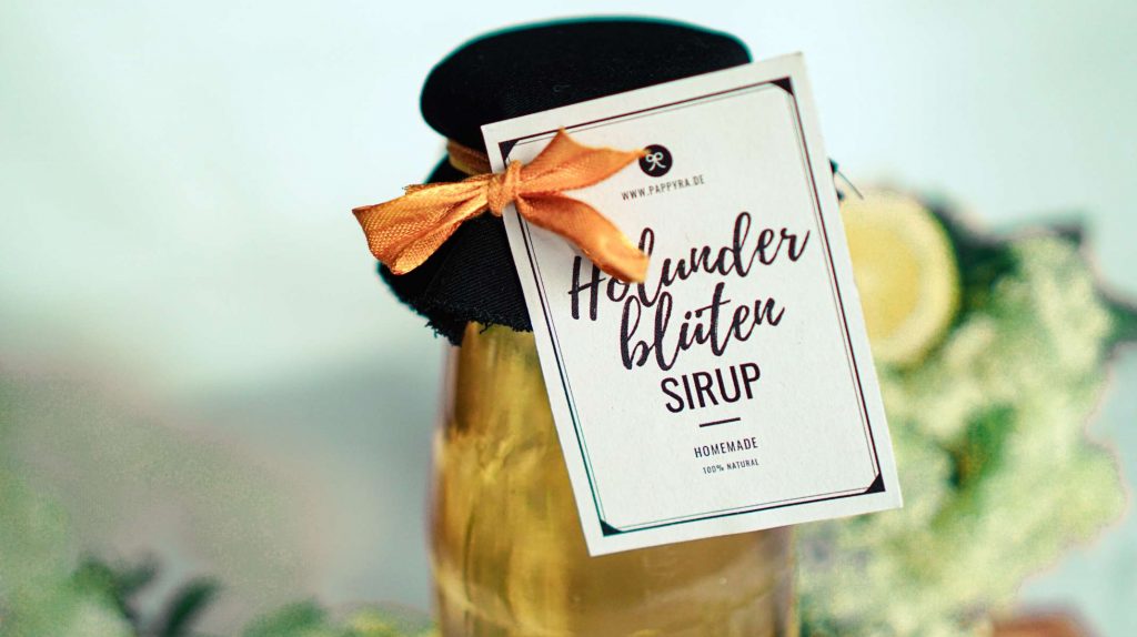 Nahaufnahme eines Holunderblütensirup Etiketts an einer Glasflasche, verziert mit Stoff und orangefarbener Schleife, im Hintergrund Holunderblüten und Zitrone.