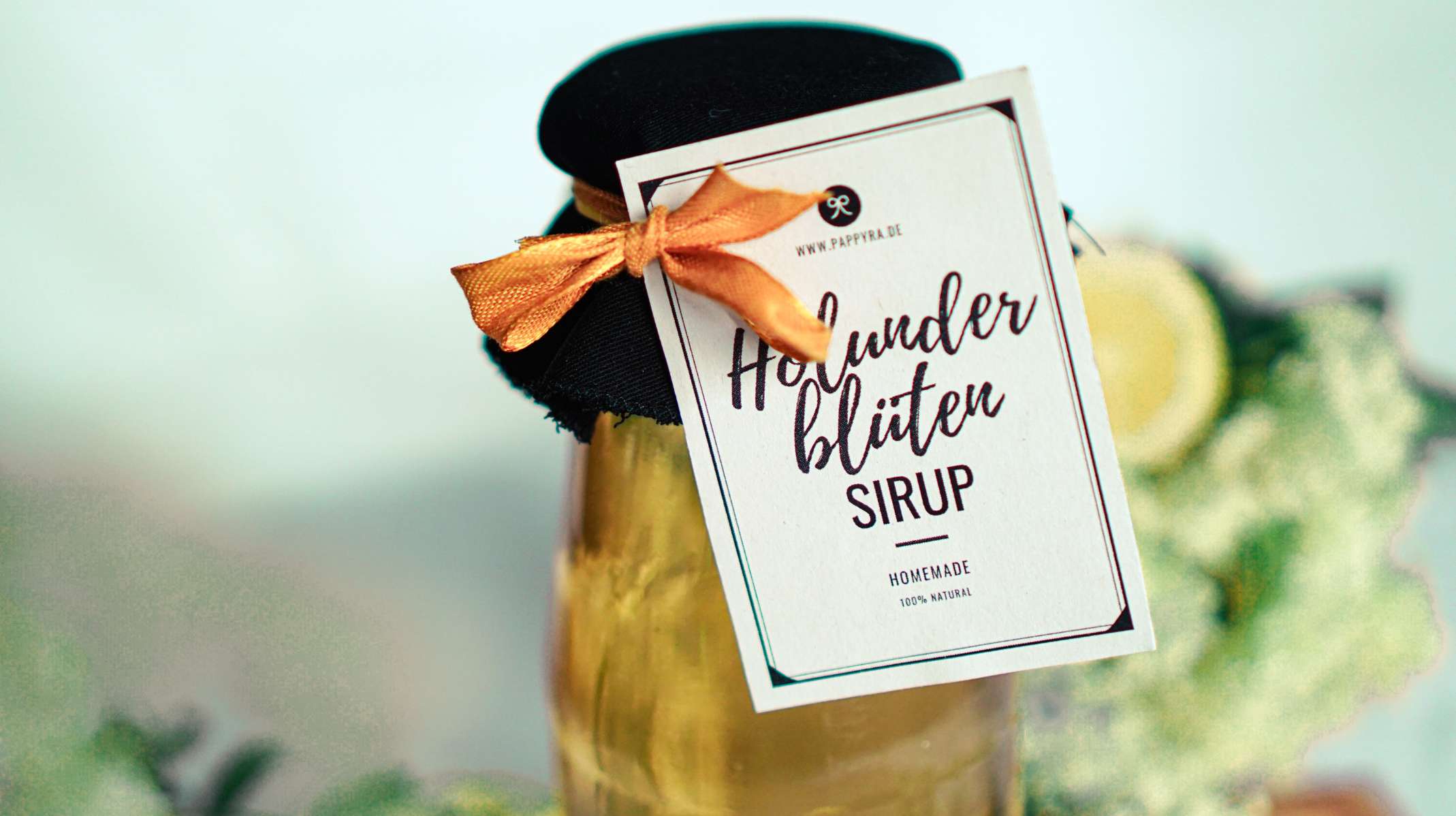 Nahaufnahme eines Holunderblütensirup Etiketts an einer Glasflasche, verziert mit Stoff und orangefarbener Schleife, im Hintergrund Holunderblüten und Zitrone.